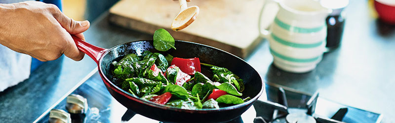 hand holding from left a red skillet with black interior filled with greens and red onions, over gas stove top burner.